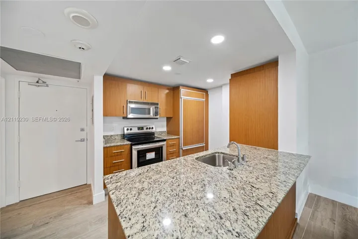 Kitchen featuring wood-finish cabinetry, stone countertops, a stainless steel undermount sink, built-in panel-ready refrigerator, and wood-finish flooring