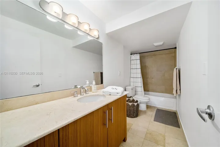 Bathroom featuring a vanity with a light-toned stone countertop, wood-finish cabinetry, a large wall-mounted mirror, four-bulb vanity lighting, and a bathtub with tile surround