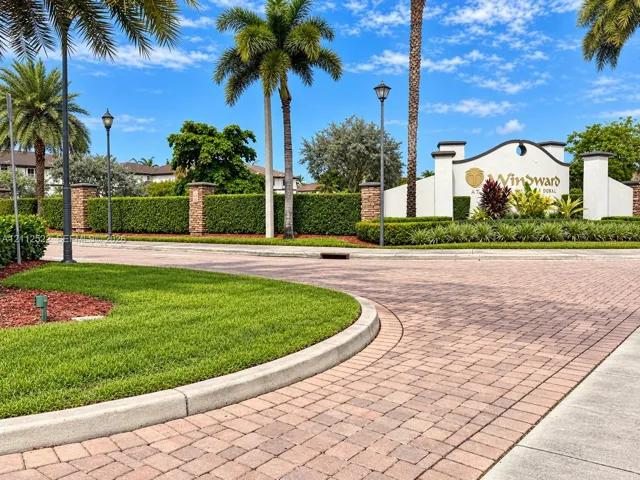 Gated community entrance featuring decorative brick paving, stone-accented masonry pillars, mature palm landscaping, and manicured hedges