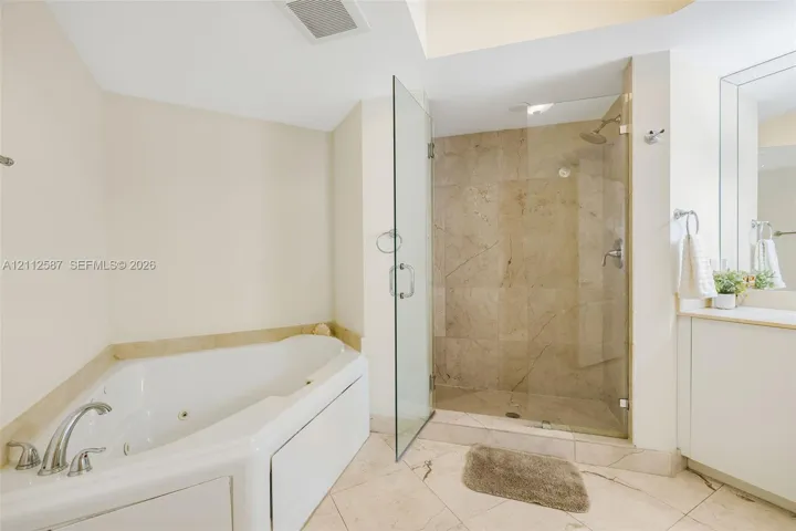 Jetted soaking tub, walk-in shower with stone-tile surround, frameless glass enclosure, stone flooring, and built-in vanity