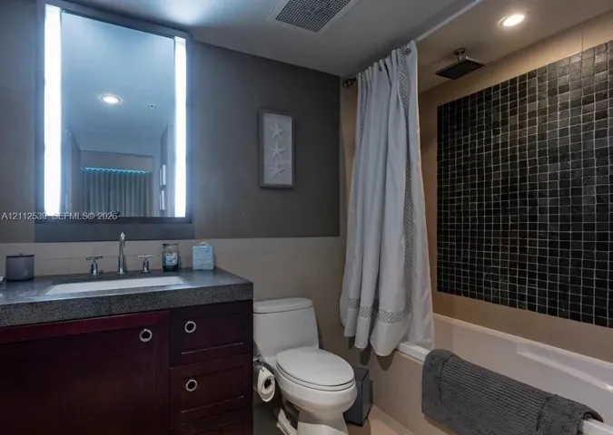 Bathroom featuring a dark wood-finish vanity with a stone countertop, an illuminated mirror, and a bathtub with black mosaic tile surround and rainfall shower head