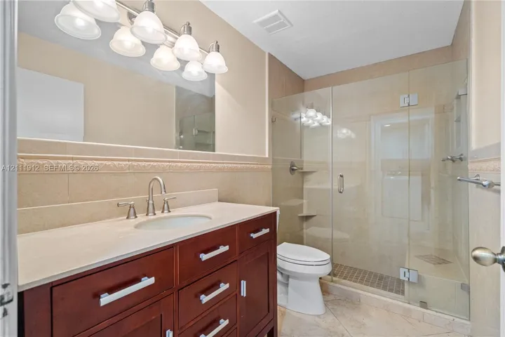 Bathroom featuring a dark wood-finish vanity, light-toned countertop with an integrated sink, frameless glass-enclosed shower, decorative tile wall border, and multi-bulb vanity lighting