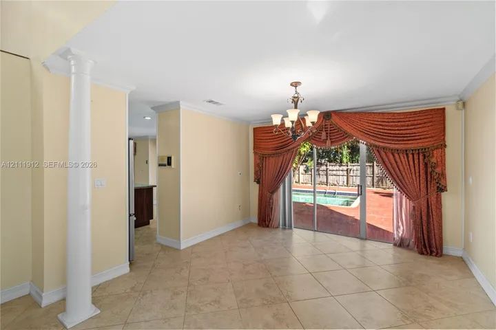Open-concept room featuring diagonal tile flooring, a decorative structural column, crown molding, a multi-arm chandelier, and sliding glass doors revealing an outdoor pool and patio