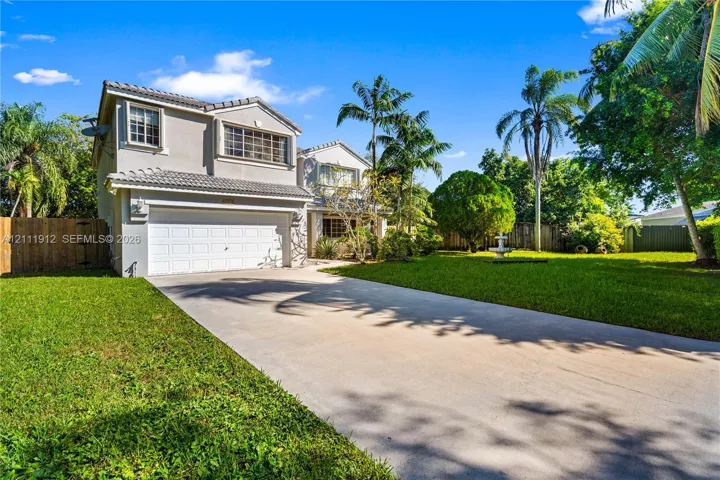 Two-story exterior featuring a light-toned stucco façade, tile roof, multi-car garage, concrete driveway, and expansive lawn with mature palm trees