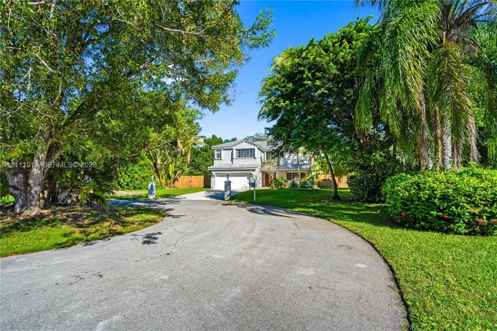 Two-story residence featuring a paved circular driveway, sprawling lawn, mature trees, and attached garage