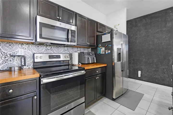 Kitchen featuring dark-finish cabinetry, butcher-block countertops, stainless steel appliances, decorative patterned backsplash, and tile flooring