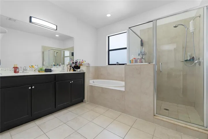 Dark cabinetry with dual undermount sinks, an expansive wall-to-wall mirror, a tiled deck-mounted soaking tub, and a separate glass-enclosed shower with tiled surround