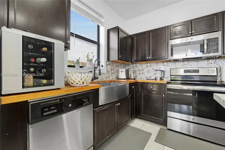 Kitchen featuring dark-finish cabinetry, butcher block countertops, patterned tile backsplash, stainless steel farmhouse-style sink, and stainless steel appliances