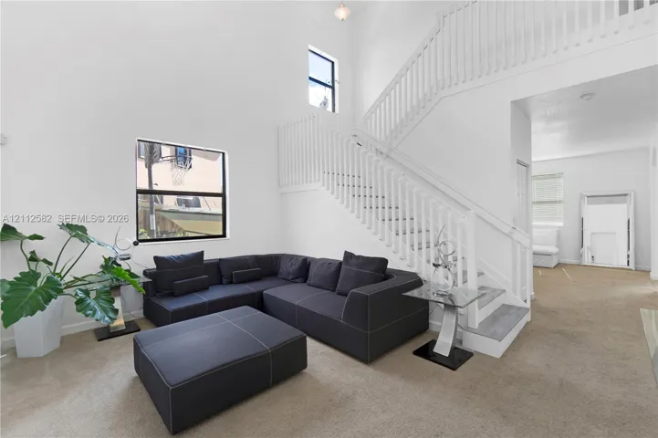 High-ceiling living area featuring a central staircase with white wooden balusters, large windows, and carpeted flooring