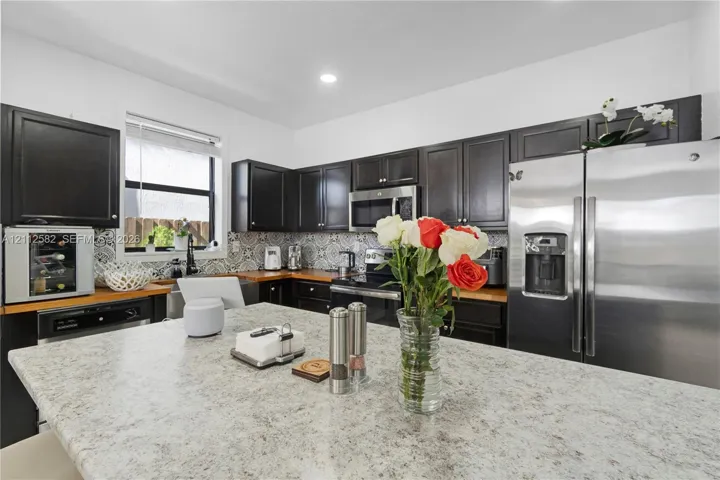 Kitchen featuring dark cabinetry, stone-surface island, patterned tile backsplash, stainless steel appliances, and wood-finish countertops