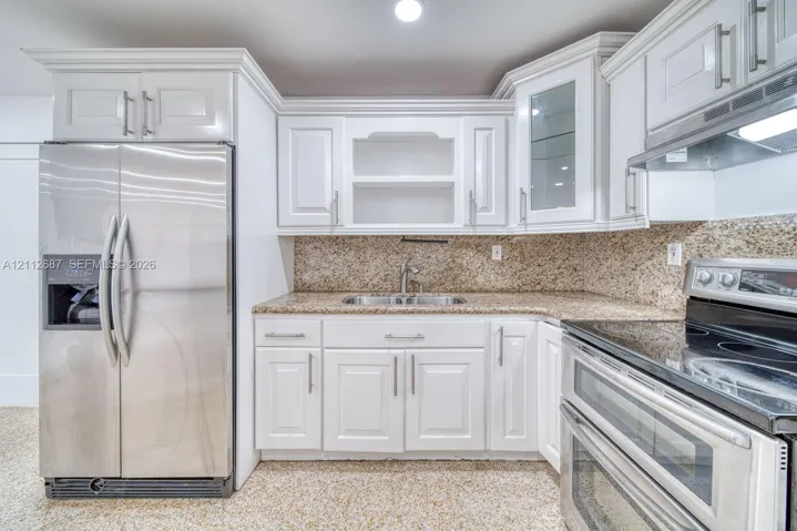 Kitchen featuring white shaker-style cabinetry, stone-finish countertops, stainless steel appliances, glass-front upper cabinet, and double-basin undermount sink