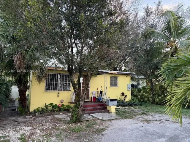 Stucco exterior with masonry construction, concrete front steps, and metal security window grilles, situated on a lot with established mature trees and palms