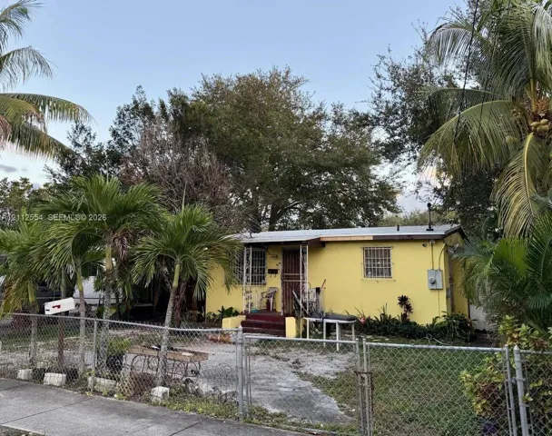 Single-story residence featuring a painted stucco exterior, metal-framed window guards, covered front entryway, and mature palm landscaping situated within a chain-link fenced perimeter