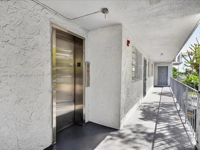 Exterior walkway featuring a brushed-metal elevator door, textured stucco wall finishes, and a metal-railing enclosure