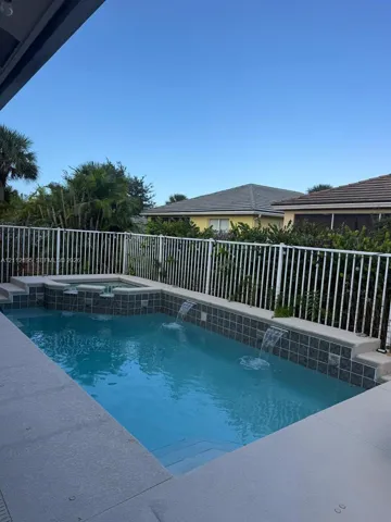 In-ground pool featuring a tiled waterline, integrated spillway spa, dual water features, and a concrete deck enclosed by metal fencing