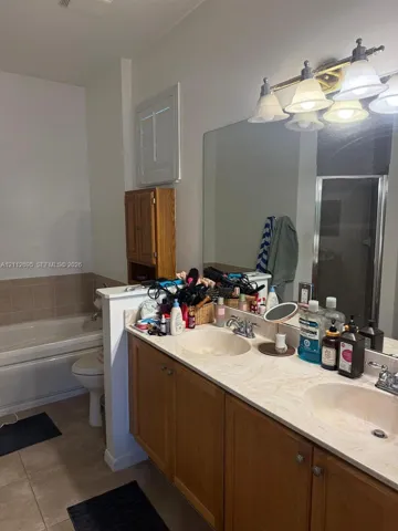 Dual-sink vanity with a solid-surface countertop and light-wood cabinetry, large wall-mounted mirror, four-light vanity fixture, built-in medicine cabinet, and a tiled soaking tub