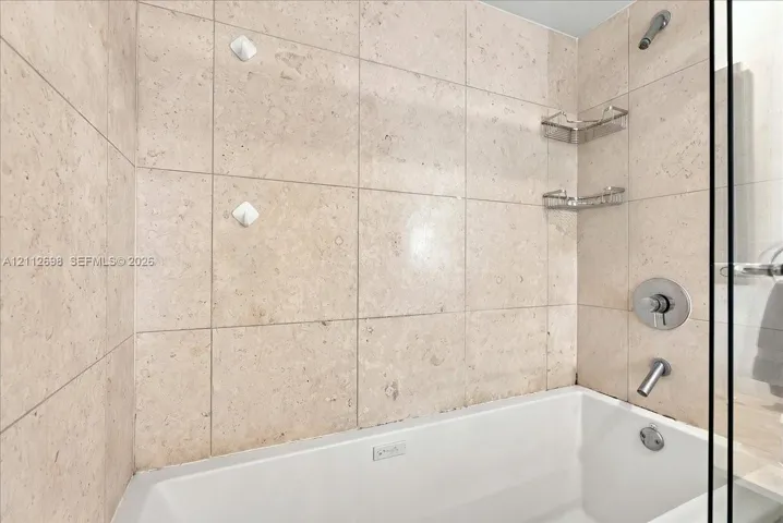 Shower and bathtub combination featuring large-format stone-finish tile surround, polished chrome fixtures, and built-in metal shelving