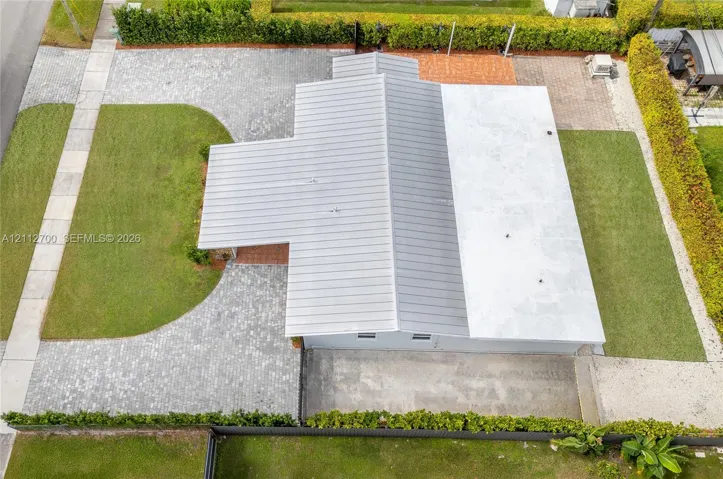 Aerial perspective featuring a metal roof, extensive paver driveway, manicured lawn, and perimeter hedging