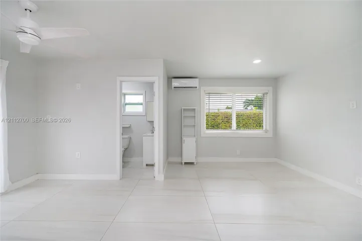 Large-format tile flooring, ceiling fan with integrated lighting, wall-mounted ductless air conditioning unit, and expansive window with horizontal blinds