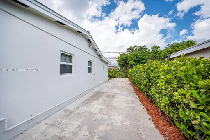 Side exterior featuring smooth stucco siding, concrete paved pathway, and mature hedgerow landscaping with organic mulch