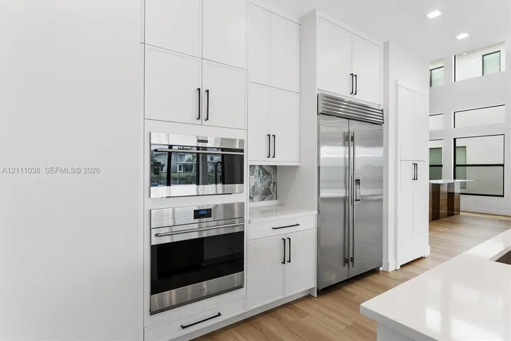 Custom floor-to-ceiling white cabinetry featuring matte black hardware, stainless steel built-in double ovens, full-size side-by-side refrigerator, and wood-finish flooring