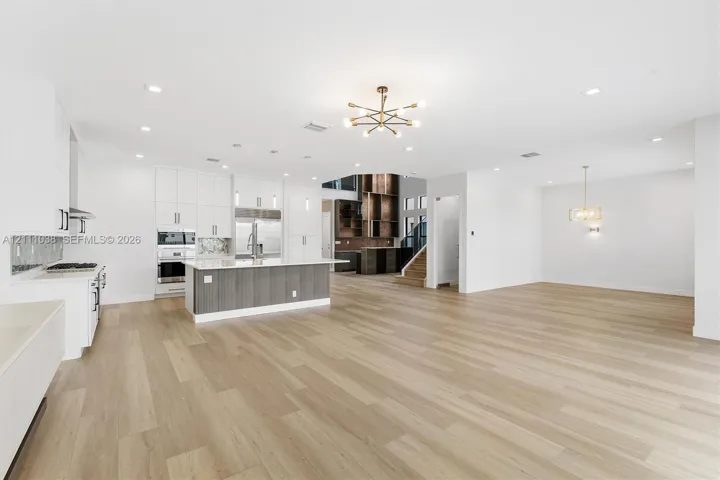 Open-concept area featuring wood-finish flooring, a kitchen island with stone countertop, white cabinetry with contrast base, modern chandelier lighting, and recessed ceiling lights