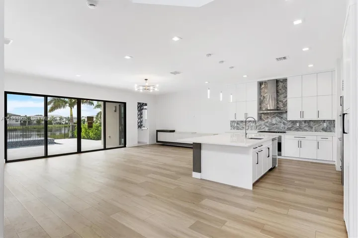 Open-concept kitchen and living area featuring wood-finish flooring, a central island with white stone countertops, floor-to-ceiling sliding glass doors, recessed lighting, and a patterned stone backsplash