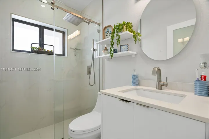 Bathroom featuring a white vanity with a neutral solid-surface countertop, walk-in shower with floor-to-ceiling tile, frameless glass shower enclosure, integrated rainfall showerhead, and circular wall-mounted mirror