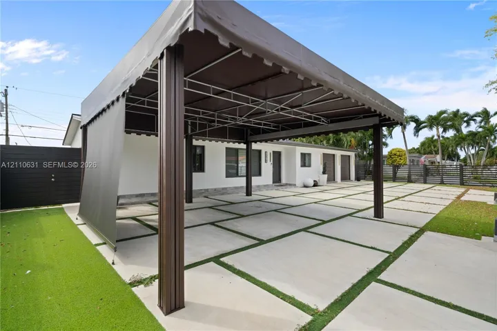 Covered metal-frame carport with integrated shade canopy, expansive concrete paver driveway featuring grass inlay, and perimeter fencing