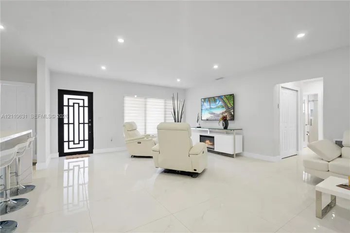 Open-concept interior featuring large-format tile flooring, recessed lighting, a modern entry door with geometric glass inlay, and a built-in media console with an integrated electric fireplace
