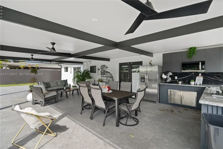 Covered outdoor patio featuring large-format tile flooring, exposed structural ceiling beams, matte black ceiling fans, and a built-in outdoor kitchen with stone-finish countertops and dark cabinetry