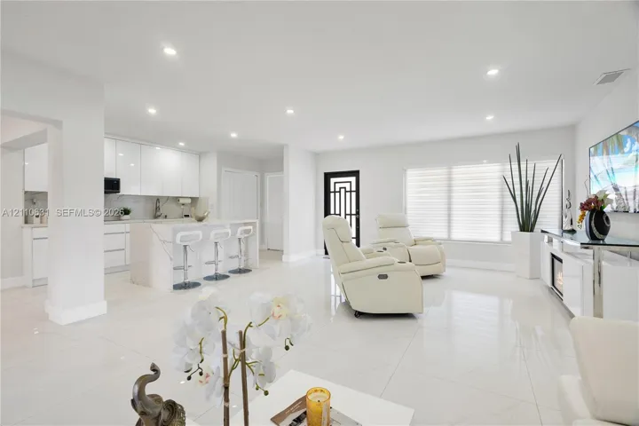 Open-concept living space featuring high-gloss cabinetry, stone-finish kitchen island with waterfall edge, recessed lighting, and large-format tile flooring