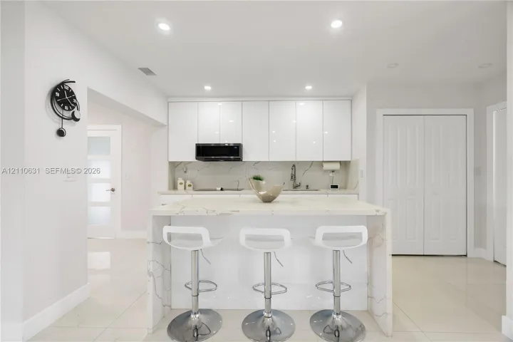 Kitchen featuring white high-gloss flat-panel cabinetry, waterfall-edge island with stone-finish countertop, recessed ceiling lighting, and tile flooring