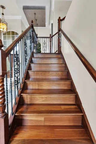 Staircase featuring wood-finish treads, decorative metal balusters, wooden handrails with carved newel posts, and a recessed ceiling detail with pendant lighting