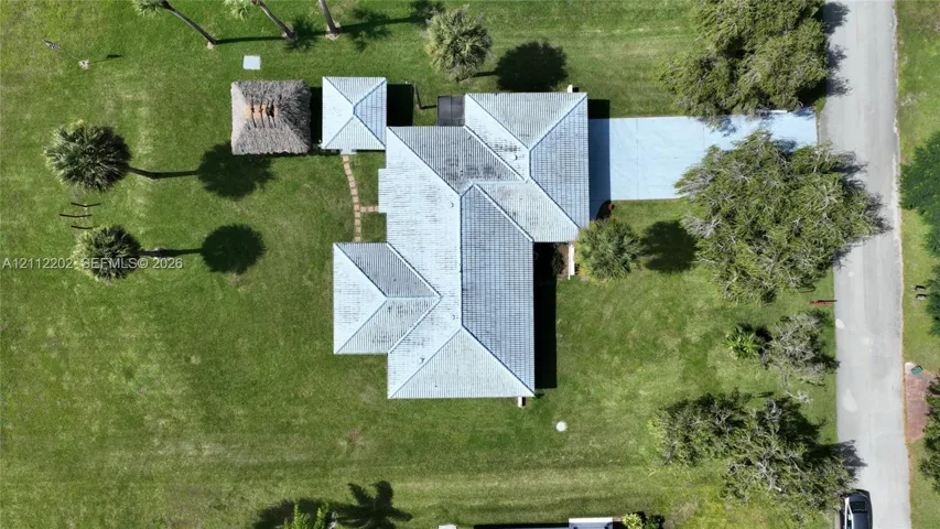 Aerial perspective of property featuring a multi-pitched metal roof, paved driveway, detached thatched-roof structure, and expansive grass lawn with mature palm trees