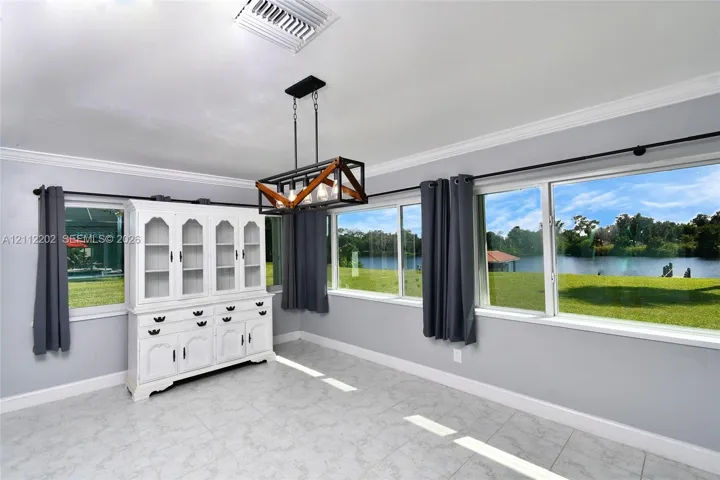 Room featuring tile flooring, crown molding, a built-in white display cabinet, a linear geometric chandelier, and expansive windows offering a water backdrop
