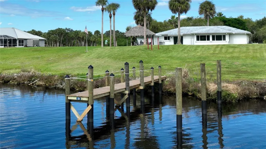 Waterfront property featuring a private wooden dock, sprawling grass grounds, mature palm trees, and a detached thatched-roof structure