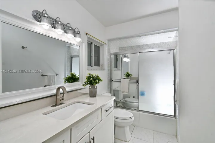 Bathroom featuring a white vanity with a composite countertop, undermount sink, wide-frame mirror, four-light vanity fixture, and a shower-tub combination with sliding glass doors