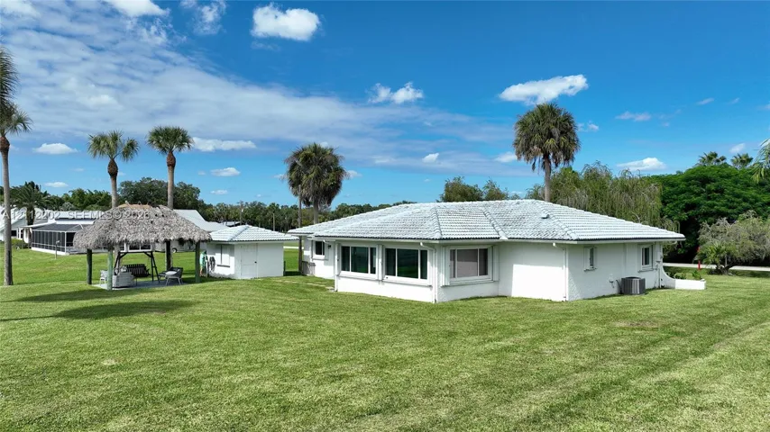 Exterior featuring white painted masonry, a hipped roof with light-colored tile, a thatched-roof structure, and an expansive lawn with mature palm trees