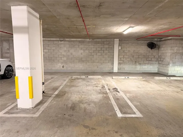 Covered parking area featuring concrete flooring, concrete block walls, painted support pillars with protective guards, and overhead lighting
