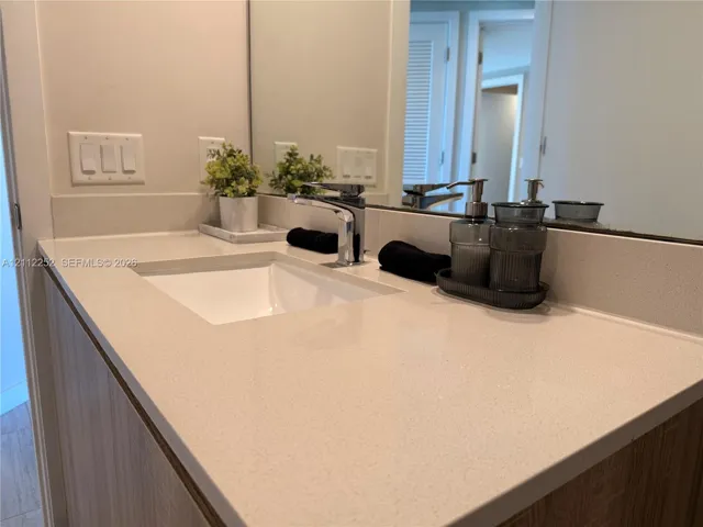 Vanity featuring a light-toned solid surface countertop, an integrated rectangular sink, a polished chrome faucet, and wood-finish cabinetry with a full-width mirror