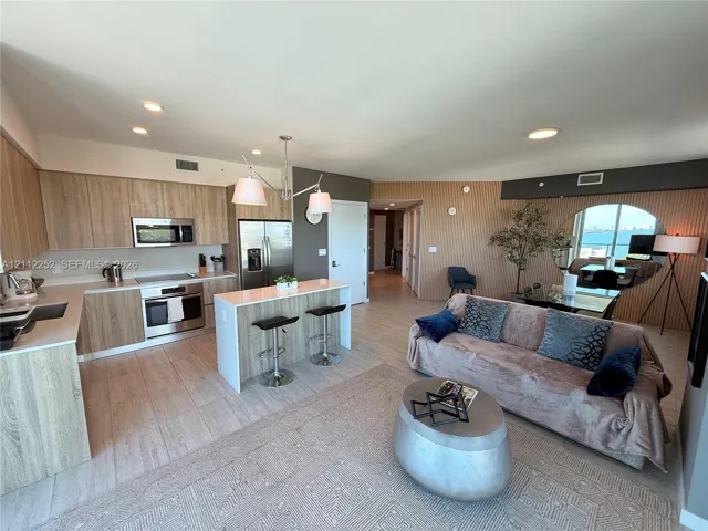 Open-concept kitchen featuring light wood-finish cabinetry, a central island with counter seating, stainless steel appliances, and wood-finish flooring throughout