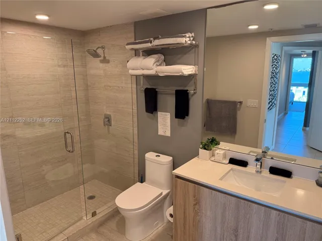 Walk-in shower with floor-to-ceiling stone-finish tile, vanity with wood-finish cabinetry, light-toned quartz countertop, integrated rectangular sink, and large wall-mounted mirror