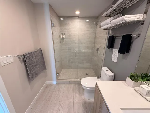 Walk-in shower with frameless glass enclosure, floor-to-ceiling stone-style wall tile, built-in shower niche, light-toned stone-style floor tile, and vanity with solid-surface countertop