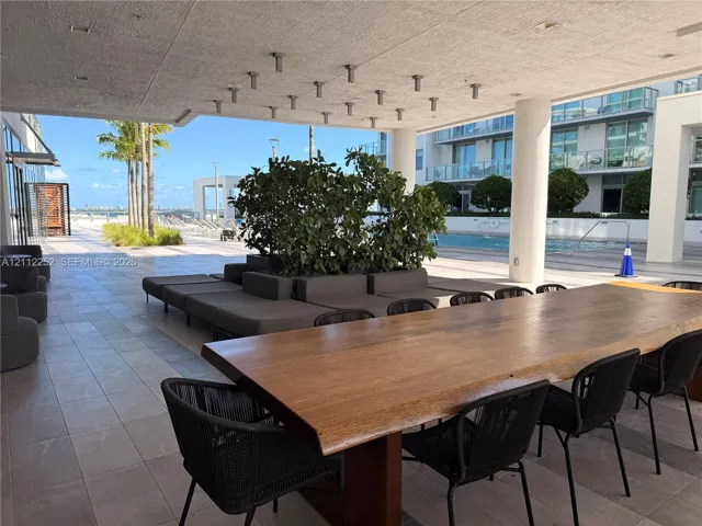Covered outdoor terrace featuring large-format tile flooring, integrated ceiling lighting, structural columns, and direct access to a communal pool area