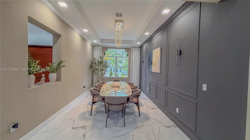 Dining area featuring large-format tile flooring, a tray ceiling with a linear crystal chandelier, an accent wall with wainscoting, and a recessed architectural wall niche