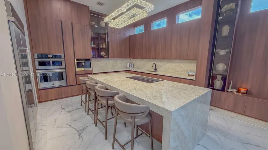 Full-height wood-finish cabinetry with integrated appliances, expansive stone-finish island, chevron-pattern tile flooring, clerestory windows, and architectural linear pendant lighting