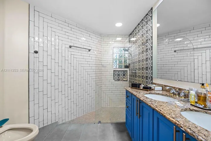 Bathroom featuring a double-sink vanity with stone-surface countertop, walk-in shower with glass panel, herringbone-patterned wall tile, and decorative accent tile work