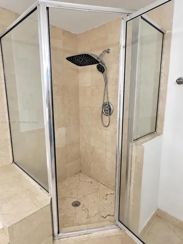 Corner shower enclosure featuring floor-to-ceiling stone-tiled walls and flooring, a built-in stone bench, a rainfall shower head with a handheld attachment, and a chrome-framed glass door