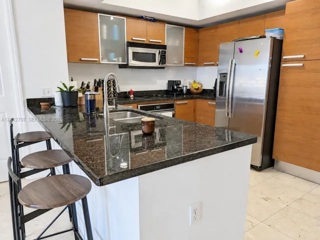 Kitchen featuring wood-finish cabinetry with frosted glass inserts, dark stone-surface countertops, a double-basin stainless steel sink with a pull-down faucet, and built-in stainless steel appliances